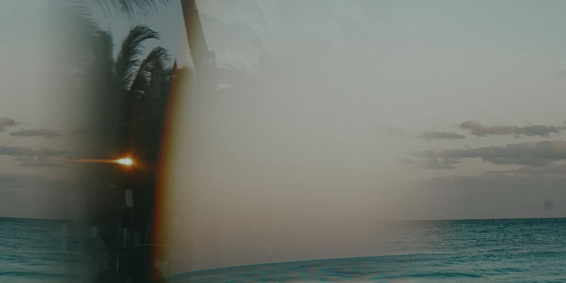 A hazy tropical seascape at dusk or dawn, with a dark palm tree silhouette on the left, a bright sun flare, and calm blue water under a pale sky.
