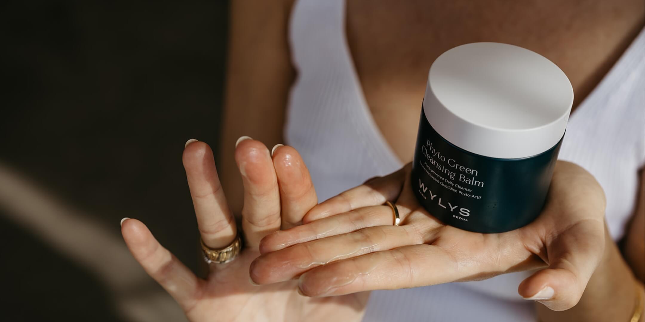 Person applying cleansing balm to hands with a container labeled 'WLYS' in the background