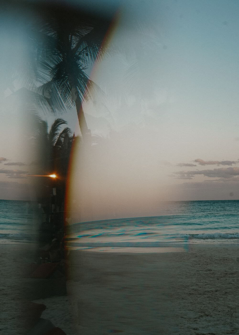 A long-exposure photograph of a tropical beach scene at sunset, featuring palm tree silhouettes, turquoise ocean waves, and a soft haze across the frame.